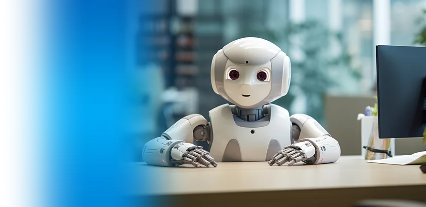 Small humanoid robot sitting at a desk beside a computer monitor